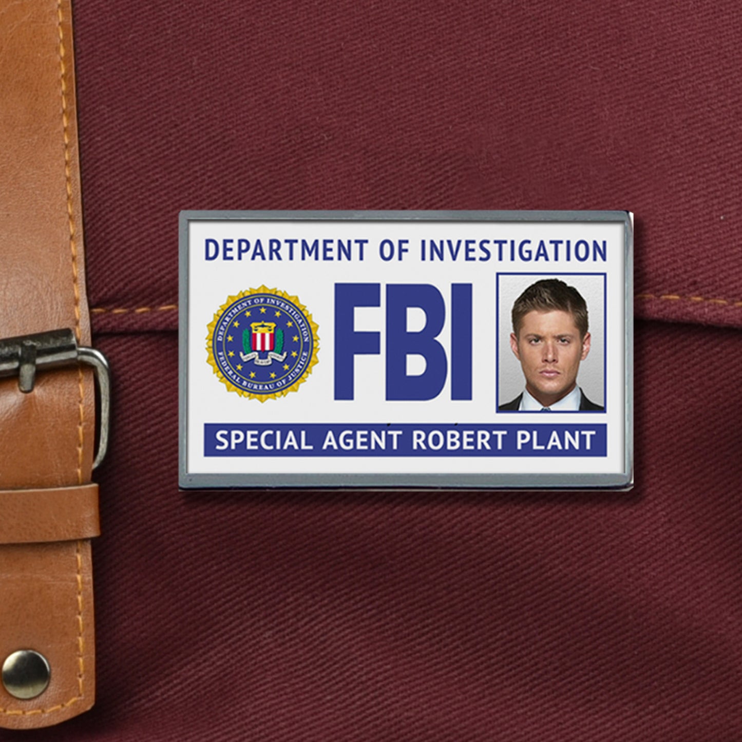 An enamel pin depicting a fake FBI badge, against a maroon canvas bag. The badge has Dean Winchester's face at the right side, with "special agent Robert Plant" across the bottom.