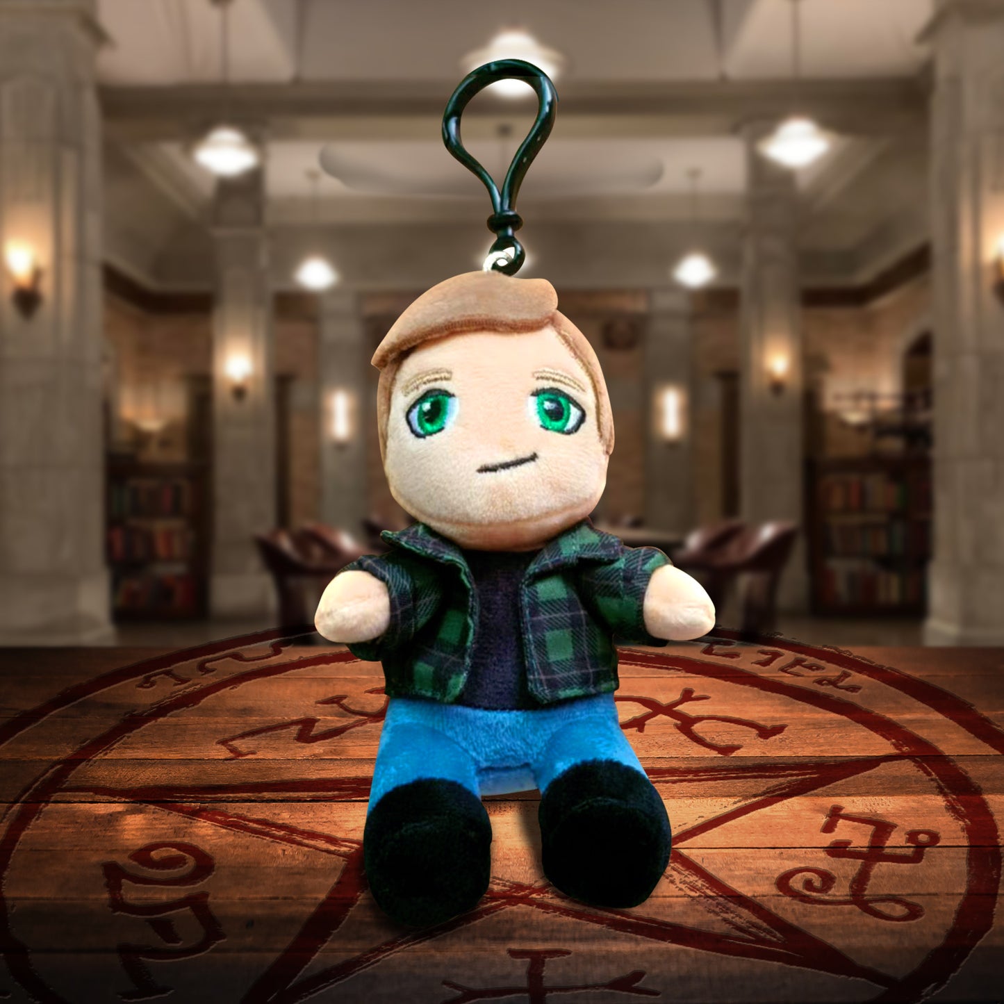 A plush keychain depicting Dean Winchester from Supernatural. Beneath the keychain is a Devil's Trap on a wooden table. In the background is The Bunker.