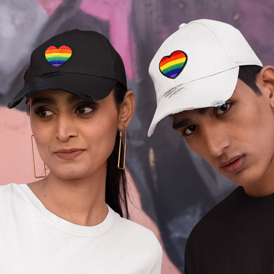 Two models side by side, in front of a painted wall. The left model is wearing a black baseball hat, the right is wearing a white baseball hat. Both hats have rainbow hearts embroidered on the front.