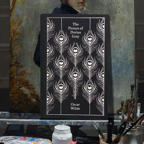 An image of a black book against a painted canvas. On the cover are white drawings of peacock feathers. White text says "The picture of dorian gray, Oscar wilde." Behind the book is a partially visible painting of a man in a blue coat. Various art supplies are standing in front of the book and canvas.