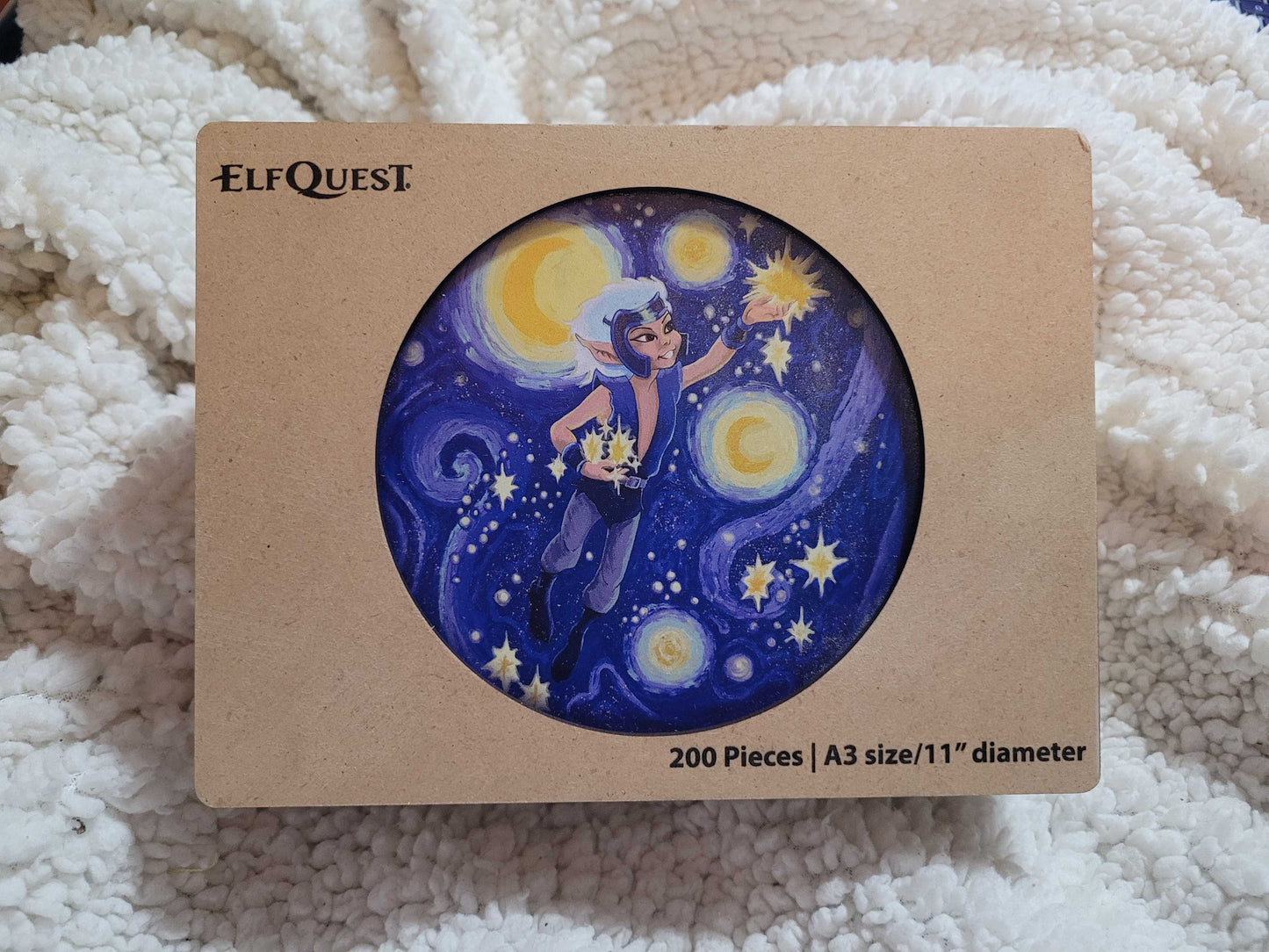 A wooden box on a white fuzzy blanket. In the center is an image depicting the ElfQuest character Skywise in the sky, gathering stars.