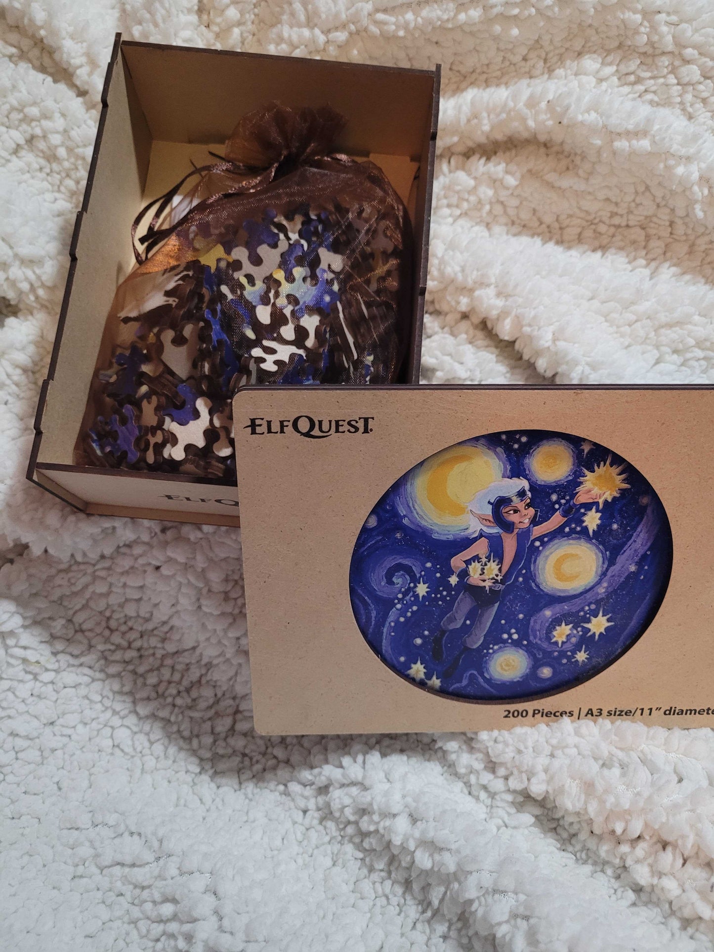 A wooden box on a white fuzzy blanket, open to show puzzle pieces inside. In the center is an image depicting the ElfQuest character Skywise in the sky, gathering stars.