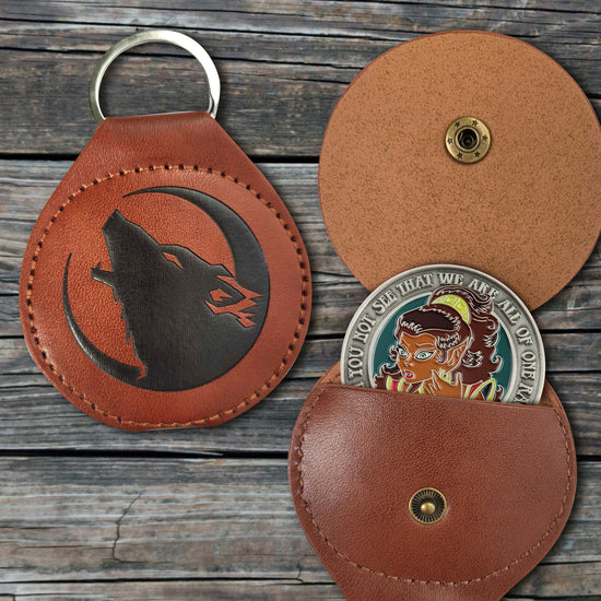 Two round, leathery coin holders with a key ring at the top. In the center of the left holder is a black embossed image of a howling wolf inside a crescent moon. The right holder is flipped open, showing an ElfQuest challenge coin inserted into the slot inside. The holders are laid against a grey wooden tabletop.