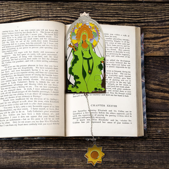 A metal bookmark sitting between pages of an open hardcover book, against a wooden table. The bookmark depicts a brown elf in a flowing green dress, with a brown headdress on her head. Behind her is a circle made up of multi-colored patchwork. A chain is attached to the bottom of the bookmark, with a yellow eight-point star charm at the end 