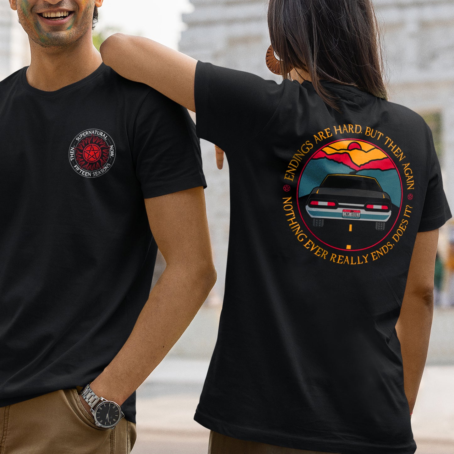 Front and back views of two models wearing a black t-shirt. On the front is the anti-possession symbol in red. Around the edge of the symbol, white text says "Supernatural, Fifteen Seasons. Then... Now..." The back of the shirt depicts the tail end of a black 1967 Chevy Impala driving on a black road toward a sunset. Around the edge is yellow text saying “Endings are hard, but then again nothing ever really ends, does it?" Behind the models is a white building.