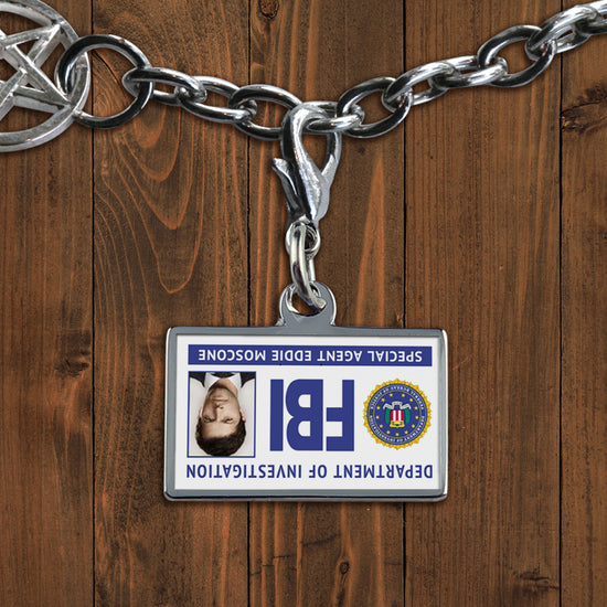 A white charm against a wooden background. The charm depicts an FBI badge, with Castiel's face at the side. At the bottom is white text saying "Special agent Eddie Moscone." The badge is upside down.