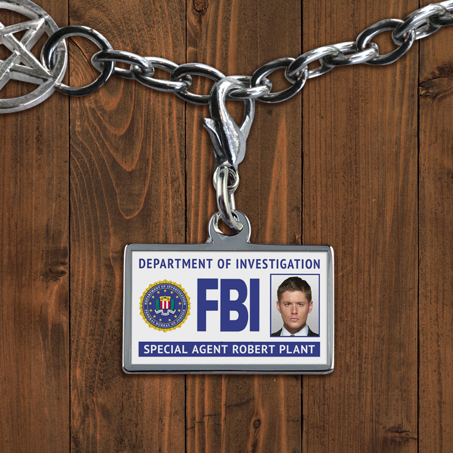 A white charm against a wooden background. The charm depicts an FBI badge, with Dean Winchester's face at the side. At the bottom is white text saying "Special agent Robert Plant."