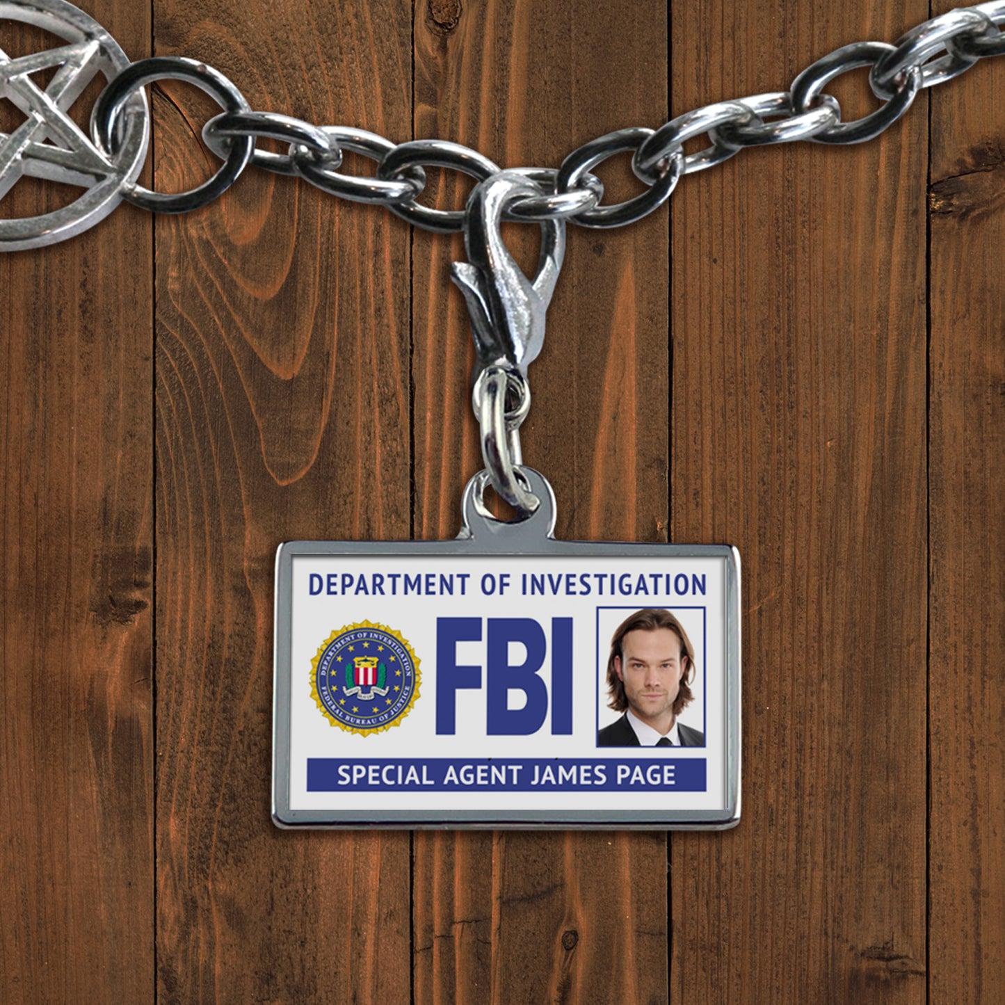 A white charm against a wooden background. The charm depicts an FBI badge, with Sam Winchester's face at the side. At the bottom is white text saying "Special agent James Page."