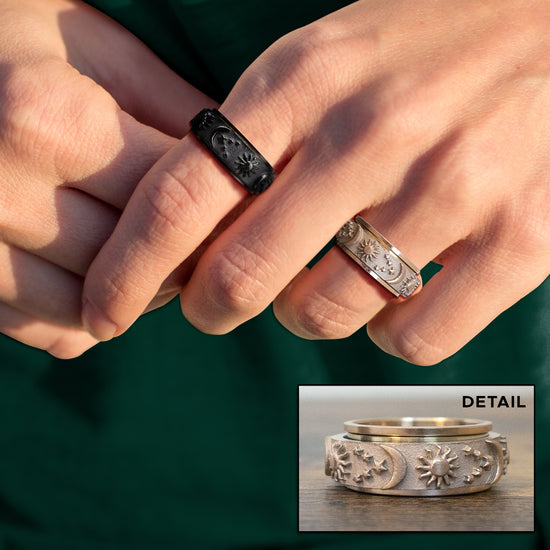 Close up of a model's hand, wearing two rings. One ring is black, the other is silver. Around the surface of the rings are raised depictions of the sun, the moon, and stars. Behind the model's hand is a dark greenbackground. At the bottom right corner is an inset image of the silver ring, showing a close up of the ring's details.