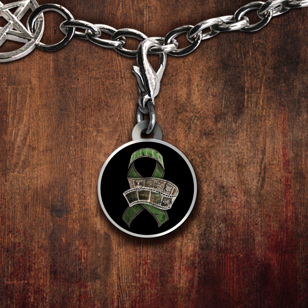 A silver-toned charm featuring a green ribbon design over a film reel motif on a black background, set against a dark wood surface.
