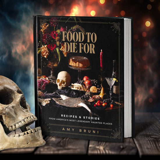 A black book on a wooden table. On the front cover of the book is a collection of berries, candles, cakes, and flowers, against a black background. Gold text at the top says "Food to die for." At the bottom is white text saying "recipes & stories from america's most legendary haunted places." Next to the book is a human skull. In the background is smoke and sparks from a fire.