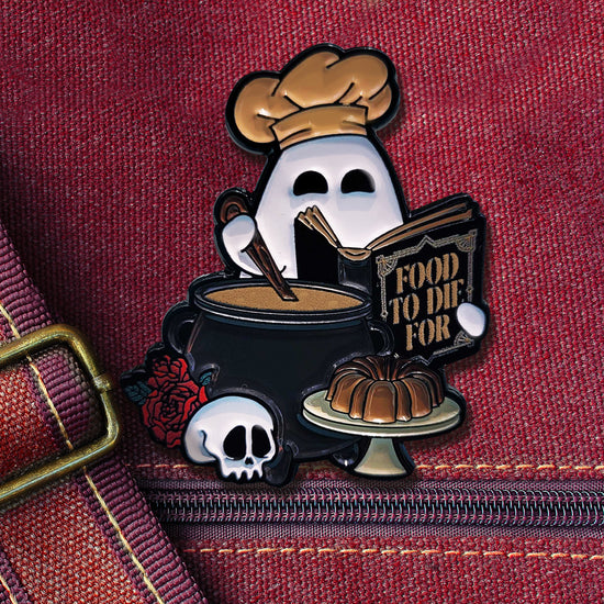 Close up view of an enamel pin on a red bag. The pin depicts a white ghost wearing a chef's hat. The ghost is holding a cookbook in one hand, and stirring a spoon in a black cauldron with the other hand. In front of the cauldron is a skull, roses, and a cake on a stand.