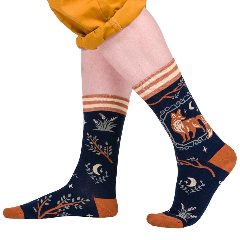 A model's feet wearing a pair of black socks. The socks feature a pattern of tree branches and crescent moons, with a brown fox on the side.