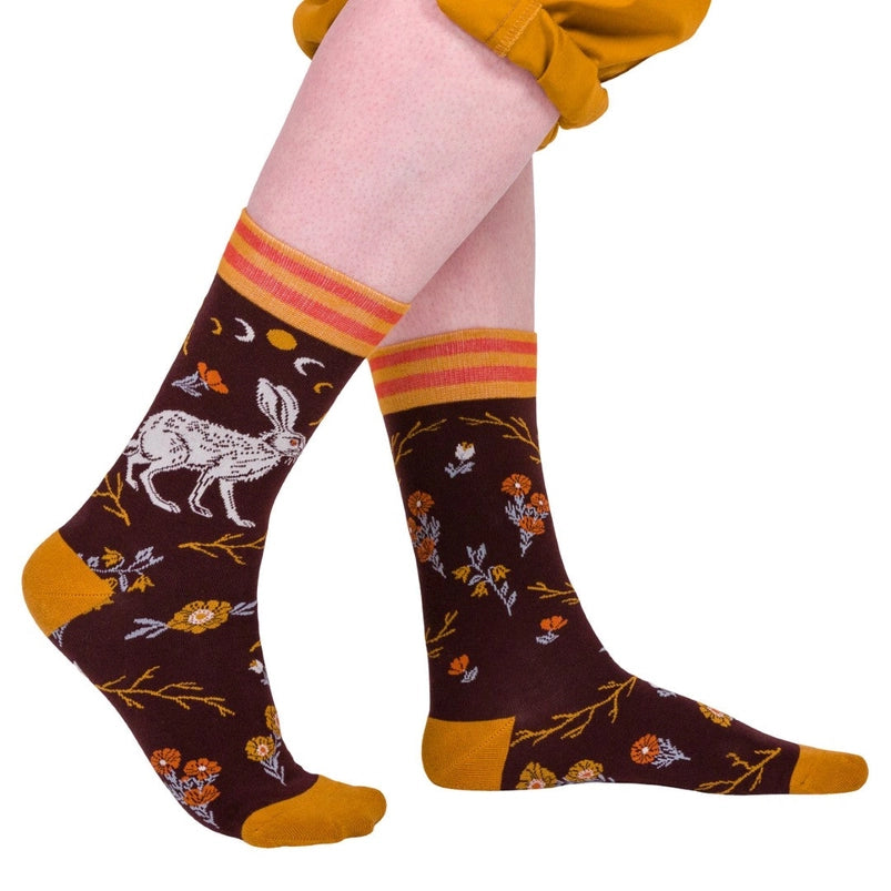 A model's feet wearing a pair of brown socks. The socks feature a pattern of red and yellow flowers, with a white hare on the side.