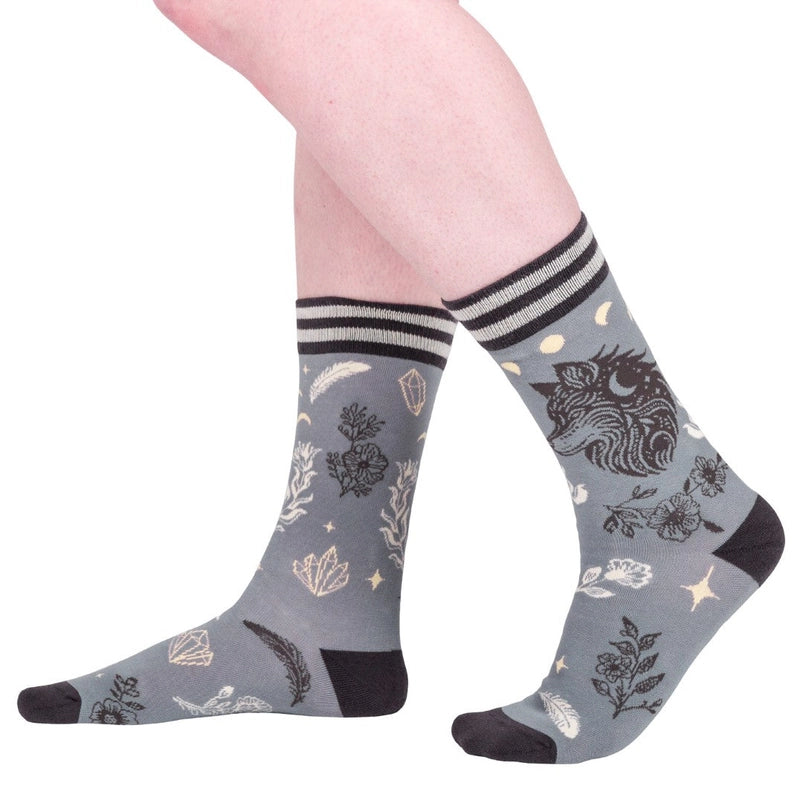 A model's feet wearing a pair of grey socks. The socks feature a pattern of white and black crystals and flowers, with a black wolf on the side.