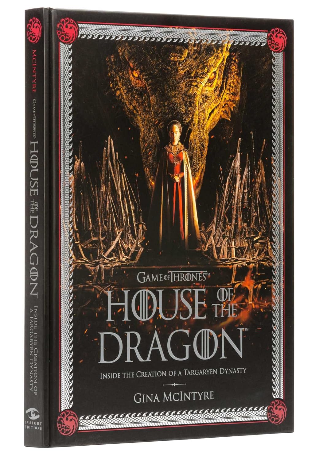 A black hardcover book against a white background. The cover depicts a woman in a red and gold dress standing on a pile of swords. with a dragon's head directly behind her. Grey text says "Game of Thrones: House of the Dragon. inside the creation of a Targaryen dynasty." Around the edge is a grey border shaped like dragon teeth, with a red multi-headed dragon silhouette in each corner.