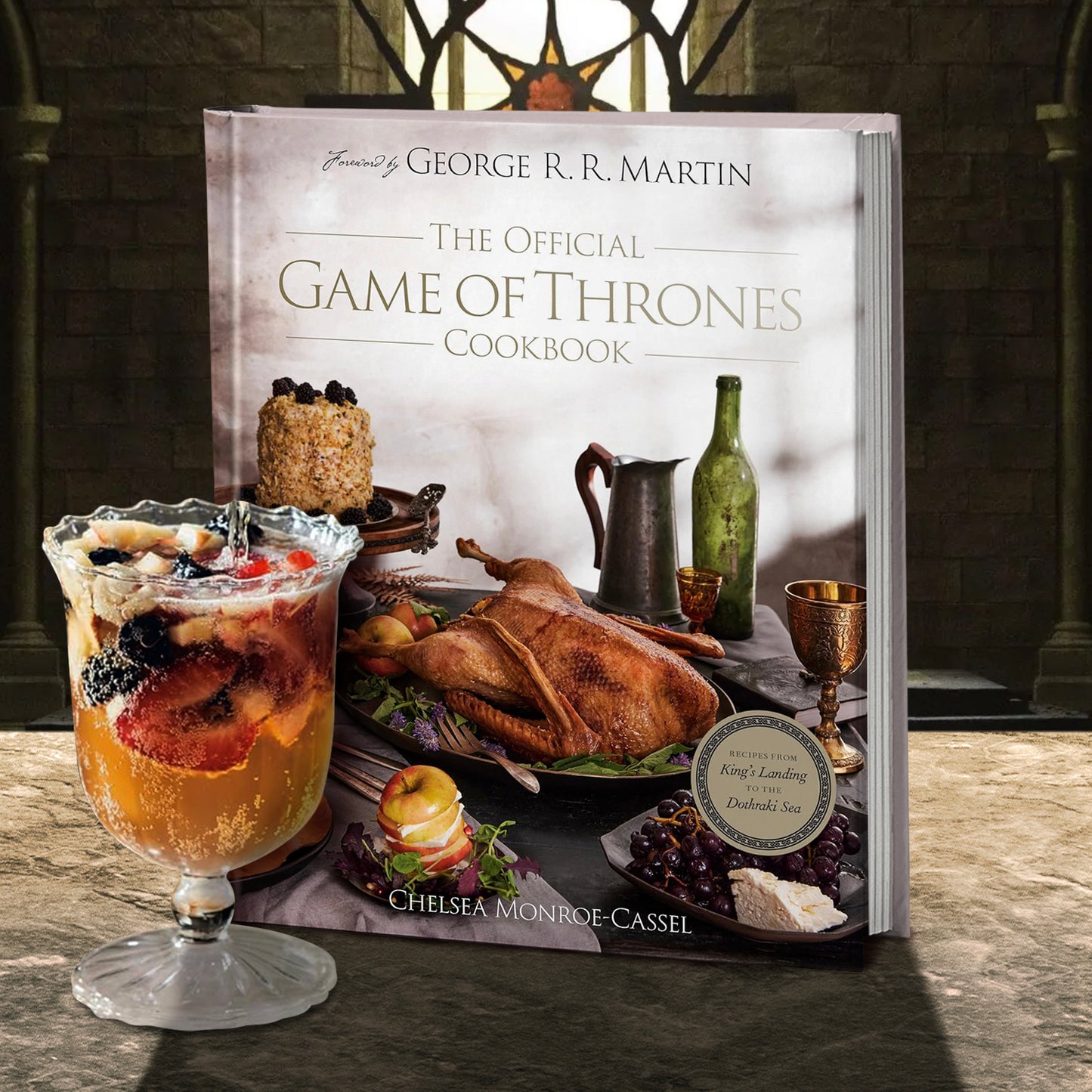 A hardcover book on a stone ledge. On the cover is a large table with various dishes from the cookbook spread across. On the side of the table is a green wine bottle and a pewter pitcher. Gold text says "The official Game of Thrones Cookbook." Next to the book is a glass goblet filled with fruit and cake. In the background is a castle room with stone walls and a stained glass window.