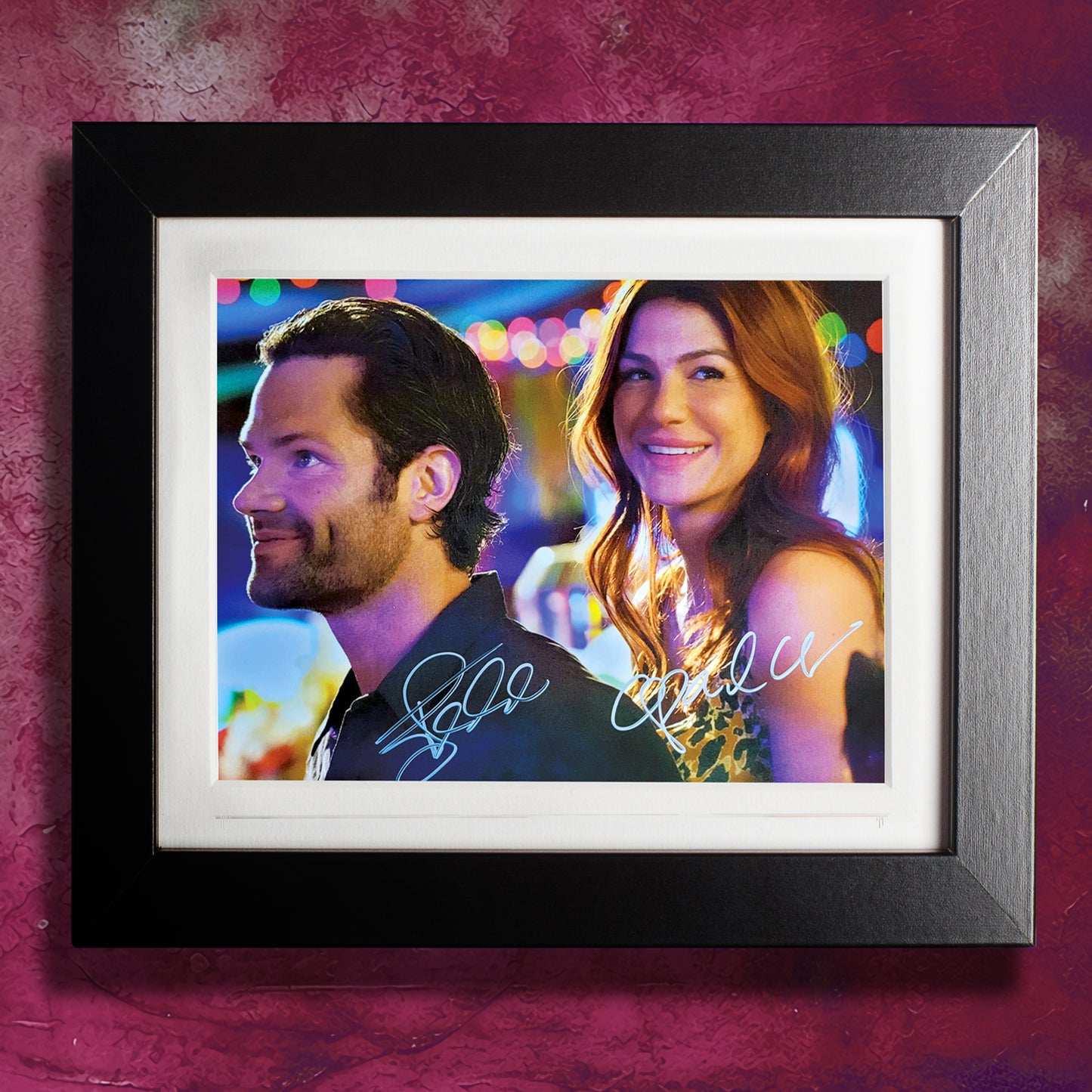 A black framed photograph against a purple-ish wall. The photograph depicts actors Jared and Genevieve Padalecki, smiling together in front of colorful lights. Beneath each of them are their autographs in white ink.