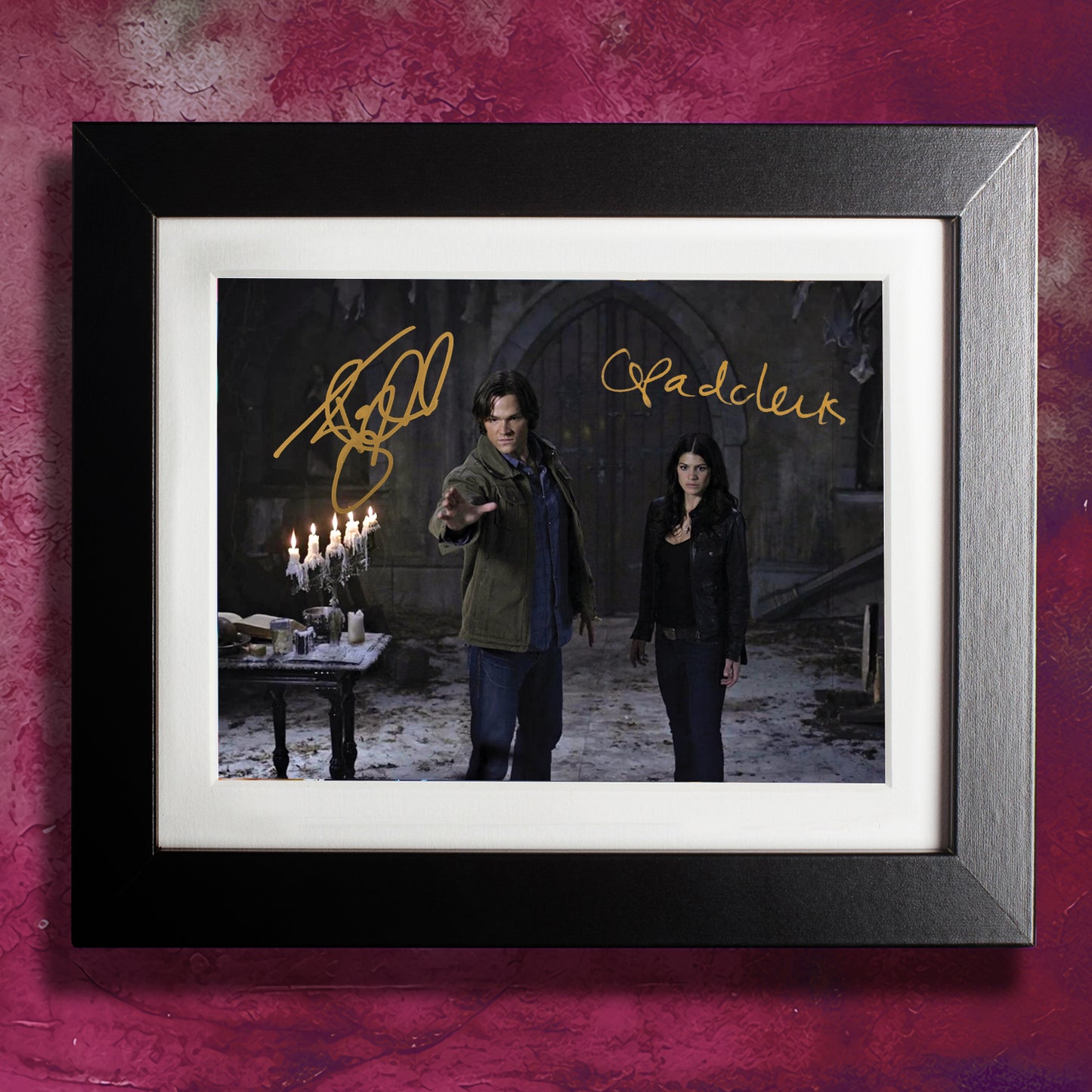 A framed picture on a red wall. The picture depicts Sam Winchester and Meg Foster in a rundown church. At the top in gold ink are Jared and Genevieve Padalecki's autographs.