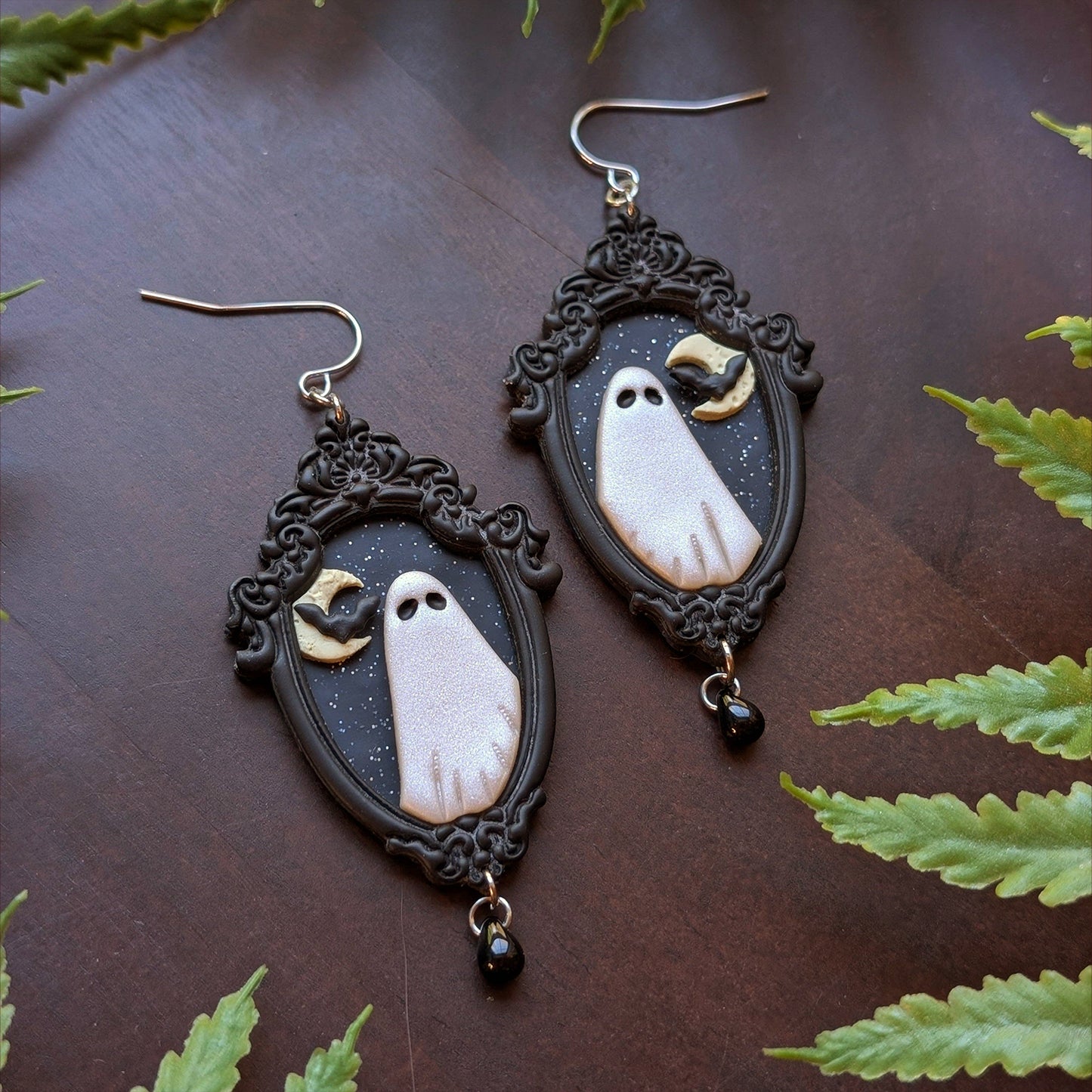 Pair of ghost-themed earrings with black frames on a dark surface with green leaves.