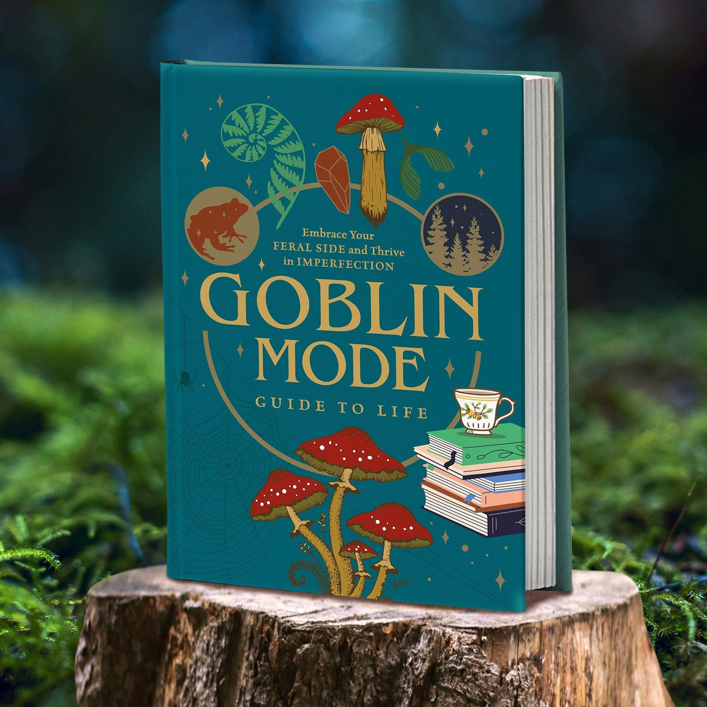 A hardcover copy of "Goblin Mode Guide to Life: Embrace your feral side and thrive in imperfection". The cover is a dark teal color with mushrooms, books, sparkles, and frogs on the front.