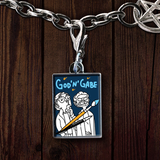 An enamel, rectangular charm on a silver chain, against a dark wood table. The charm has a blue background, and has a black and white line drawing of two male characters. One is wearing glasses and drinking from a can through a straw. The other has a scraggly beard and is scowling. Above them are drawings of a pencil and a paintbrush.