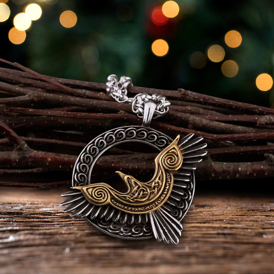 Close up of a silver and brass necklace with a silver chain, on a wooden plank. The pendant is a silver circle, empty in the center, with a Norse pattern etched around the edge. At the bottom is a gold and silver raven, wings outstretched. Behind the pendant is a bundle of twigs, with colored lights in the background.