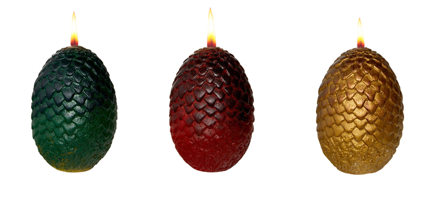 Three egg-shaped candles, with scales on their outsides, side by side. The left egg is green, the center is red, and the right is gold. Each has a burning wick at the top.