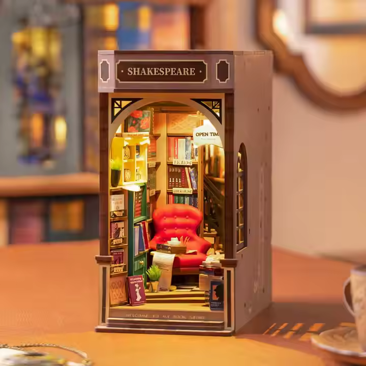 An assembled wooden book nook light, depicting Shakespeare's Bookstore in France, on a shelf. The interior is filled with tiny books on shelves, surrounding a red leather armchair and a table with a miniature teacup and saucer on it. On the front of the model is a black sign with "Shakespeare" in gold text. 