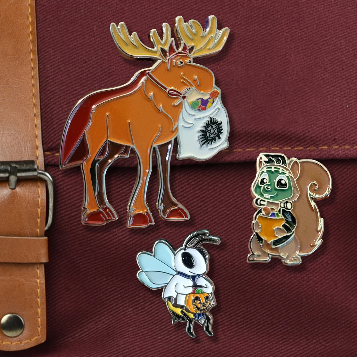 A closeup on a red bag with metal buckle. There are three pins on the bag, one large moose in a devil costume carrying a sack full of candy with an anti-possession symbol on it, one medium-sized squirrel with green Frankenstein face paint and neck bolts holding an acorn full of candy. There is a small skeleton handprint on his left shoulder. The last pin is a small bee wearing a ghost costume. He is wearing a backwards blue tie and holding a jack-o-lantern full of candy.