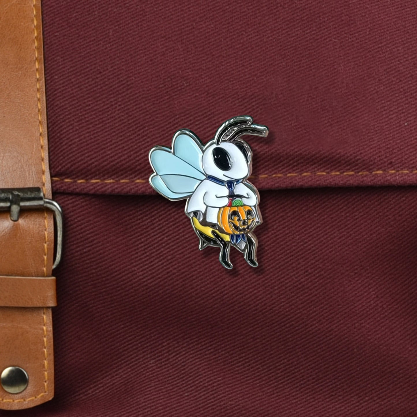 A closeup on a red bag with metal buckle. There is a small pin affixed to the bag. It is a bee wearing a ghost costume. He is wearing a backwards blue tie and holding a jack-o-lantern full of candy.