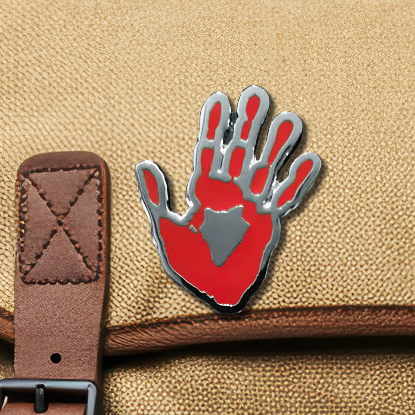 A red and silver enamel pin on a light tan canvas bag. The pin depicts Castiel's handprint.