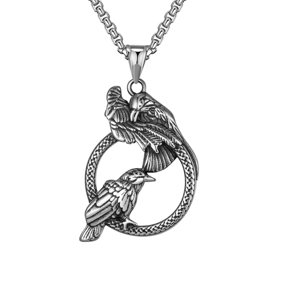 Close up view of a stainless steel pendant against a white background. The pendent depicts two ravens sitting on a ring. The top raven's wing is lifted. Attached to the pendant is a stainless steel chain.