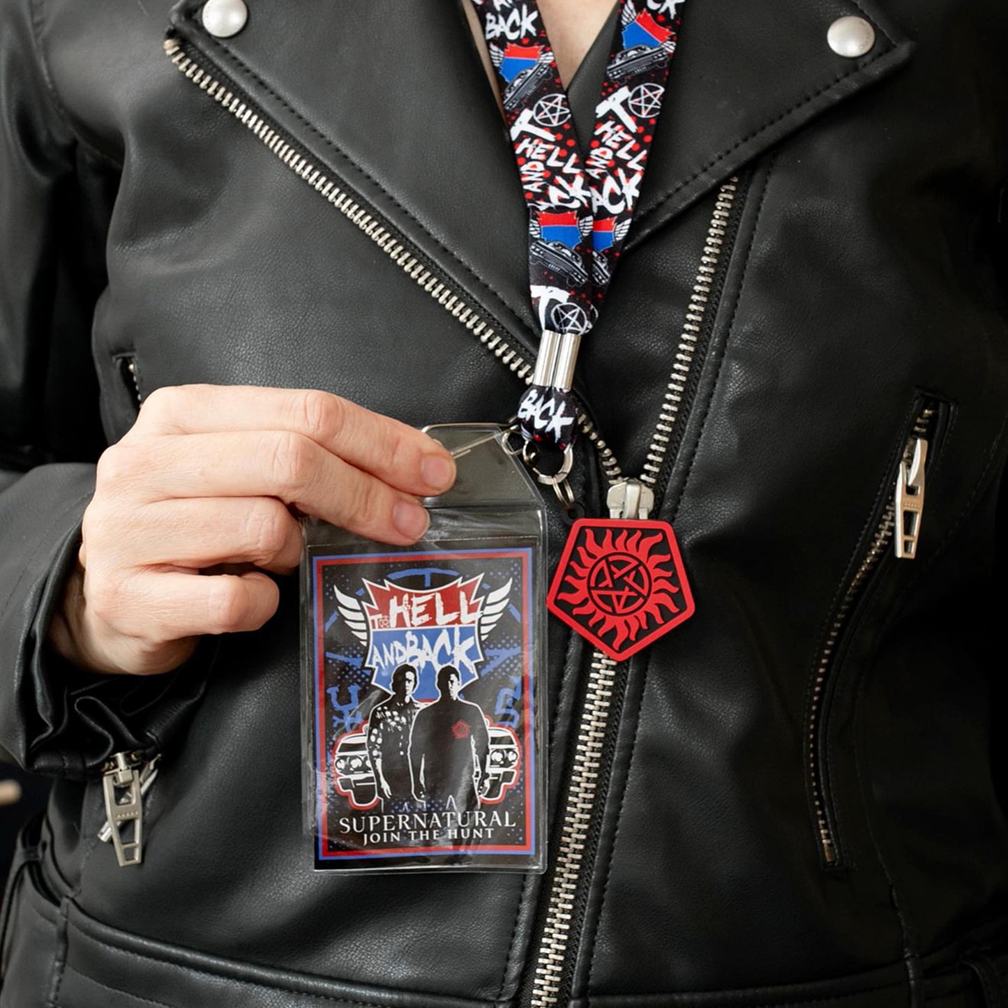 A model in a black leather jacket, wearing a lanyard with an anti-possession charm in red and black attached to the end. The lanyard features "to hell and back" in white text. Next to the lanyard is a badge featuring Sam and Dean Winchester. A red and blue road symbol at the top has white text saying to hell and back.