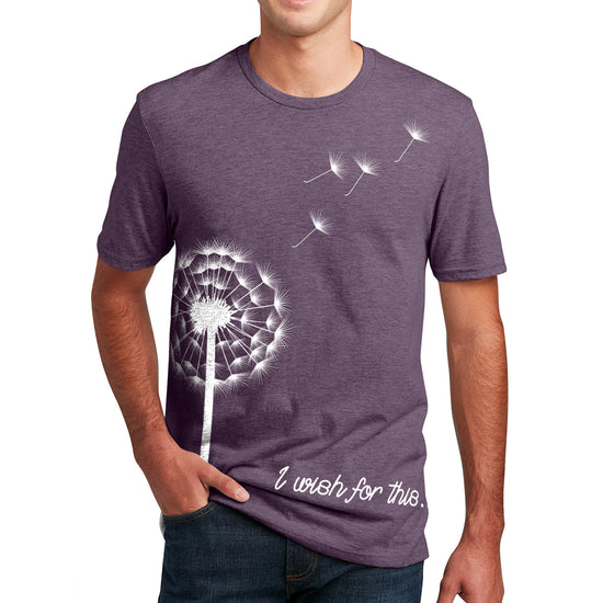 A male model wearing a purple t-shirt. On the front is a white drawing of a dandelion with four seeds floating off. At the bottom is white scripted text that says I wish for this.