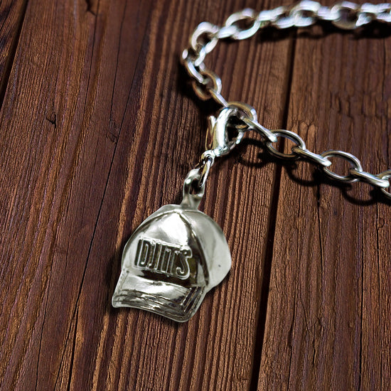 A brass charm in the shape of a baseball cap, against a white background. On the front of the cap are raised letters saying "idjits." Behind the charm is a wood background.