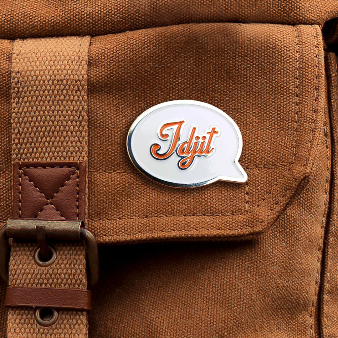 A silver pin with a white front in the shape of a speech bubble, attached to a brown canvas bag. The word "Idjit" is written in orange script font.