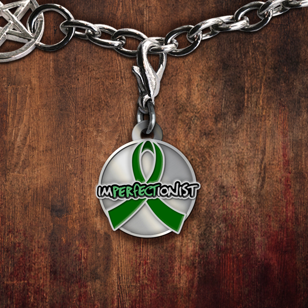 A silver-toned round charm featuring a green awareness ribbon and the word 'IMPERFECTONIST' in black outlined lettering, shown hanging from a silver chain against a dark brown wood background.