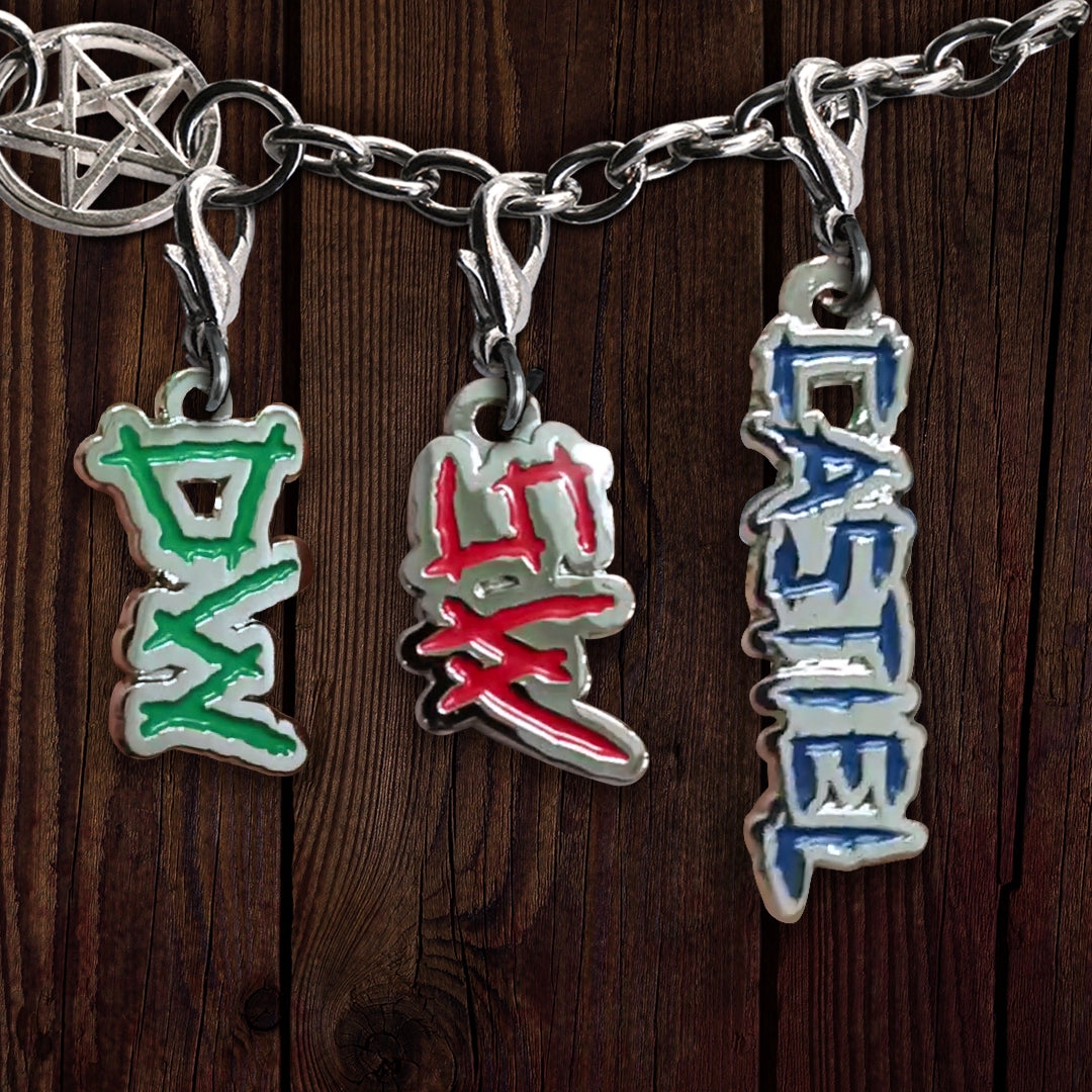 Three metal charms on a liked bracelet, against a dark wooden background. The left charm depicts the initials DW in green, the center depicts SW in red, and the right depicts the name Castiel in blue. At the top left is a charm with the anti-possession symbol at its center.