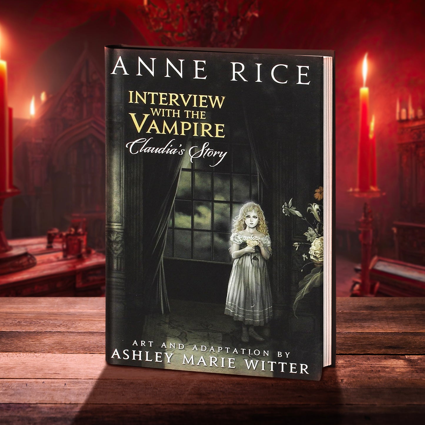 A black hardcover book on a wooden table, in front of a red-tinged gothic room with red candles. On the front cover of the book is a young girl in an old-fashioned white dress, standing in front of a window showing the nighttime sky. White and yellow text says "Anne Rice, Interview with the Vampire, Claudia's Story. Art and adaptation by Ashley Marie Witter."