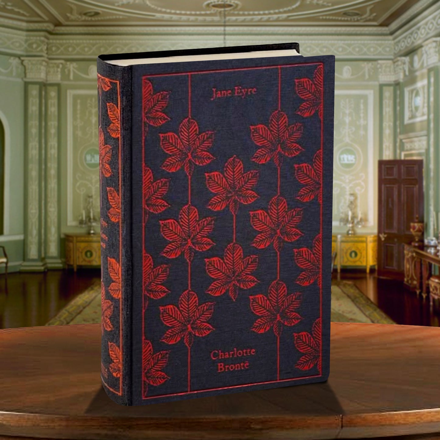 S black and red book on a wooden table. On the cover is a repeating pattern of a red leaf. Red text says "Jane Eyre. Charlotte Bronte." Behind the book is an elegant parlor room.