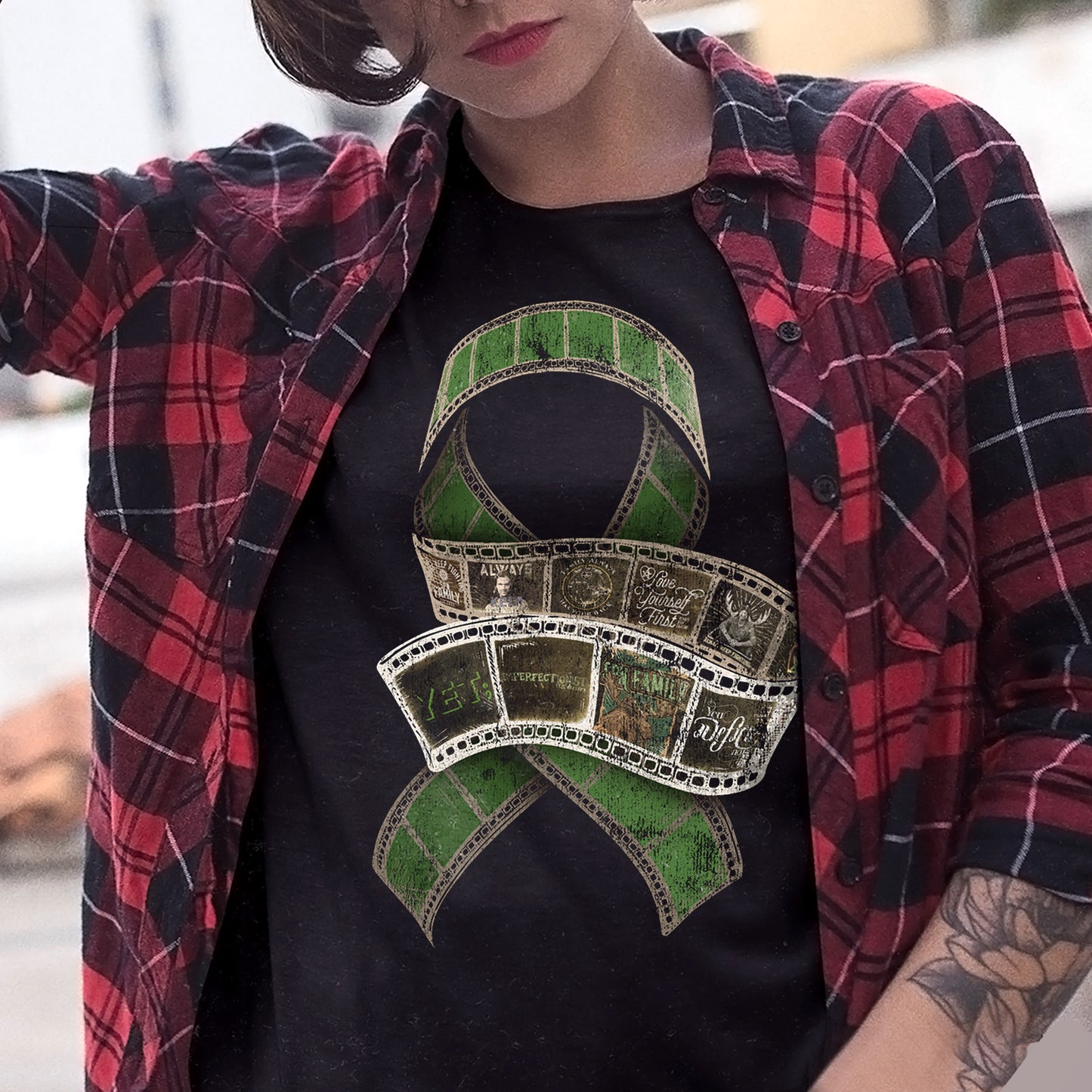 A model wearing a black T-shirt under a red and black plaid shirt. On the front of the t-shirt is a large ribbon made of film cels. In front of the ribbon is a coiled film strip with images of various Always Keep Fighting designs in the cels. Inset at the bottom left corner is a close up of the sleeve, featuring a large green X with AKF in white letters on top of it.