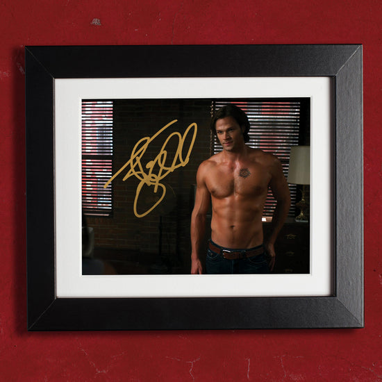 A framed picture on a red wall. The picture depicts Jared Padalecki as a shirtless Sam Winchester. Inside the image is Jared Padalecki's autograph in gold ink.