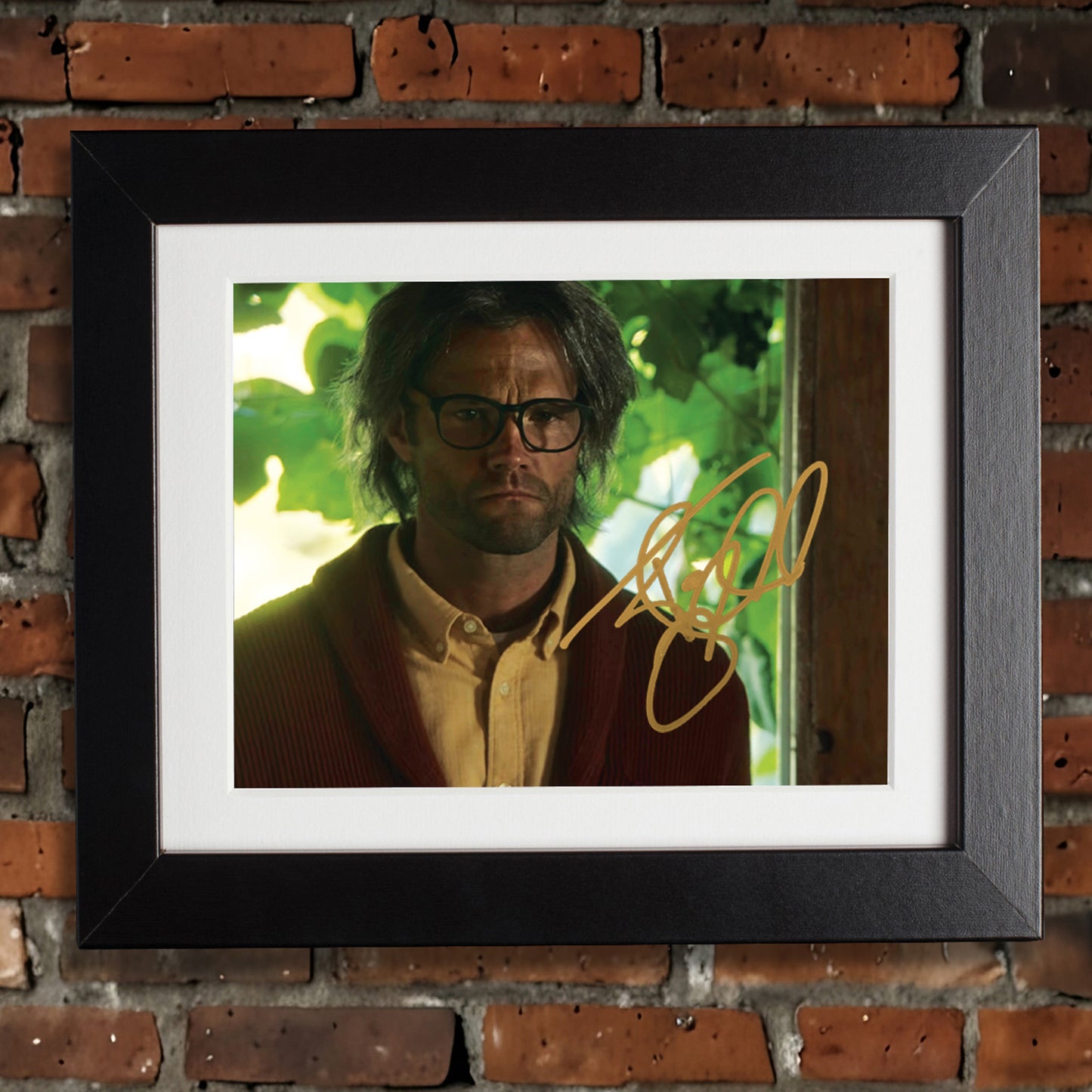 A framed picture on a brick wall. The picture depicts Jared Padalecki as an older Sam Winchester. Inside the image is Jared Padalecki's autograph in gold ink.