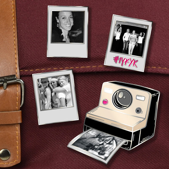 A close-up of 4 enamel pins on a maroon canvas bag. The top left pin is in the shape of a white polaroid containing a black-and-white photo of Jennifer Beals. The top right is a polaroid depicting Katherine Moennig, Leisha Hailey, Laurel Holloman, and Erin Daniels with a hot pink polaroid 'caption' reading #IYKYK. The bottom left is a polaroid depicting a group hug between Rachel Shelley, Pam Grier, and Laurel Holloman. The bottom right is a cream-and-black Polaroid camera printing a 'Bette + Tina' photo.