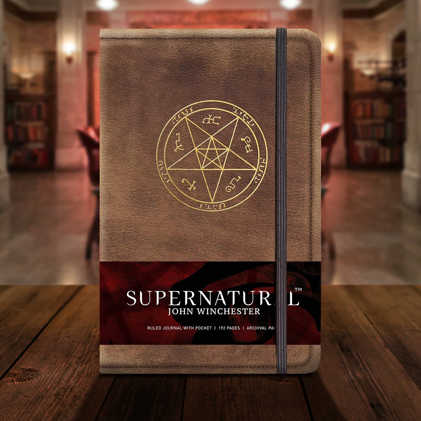 A brown leather-textured hardcover journal with a thin black strap over the right side. The journal features a gold devil's trap sigil in the center. The packaging strip reads "Supernatural: John Winchester - Ruled Journal with Pocket - 192 Pages - Archival Paper"