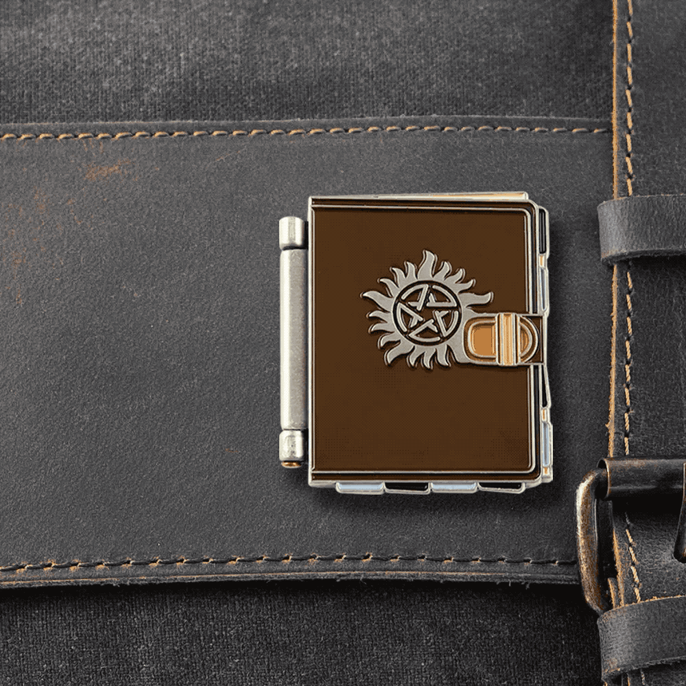 Close up image of a brown enamel pin attached to a black canvas bag. The pin is in the shape of a journal, with the anti-possession symbol on the cover in silver. The book opens to reveal two white pages, with various symbols drawn on them, and a drawing of a monster in the top left corner.