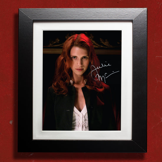 A black framed photograph against a red wall. The photograph depicts actor Julie McNiven, dressed as Anna Milton from the series "Supernatural." Julie is in a white shirt and black jacket, staring forward menacingly. On the top right side is Julie's autograph in white ink.