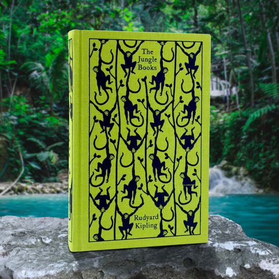 A bright green book with a pattern of monkeys drawn across the cover. Text at the top says "The Jungle Books" Rudyard Kipling, The book is resting on a grey rock, with a jungle and river in the background.