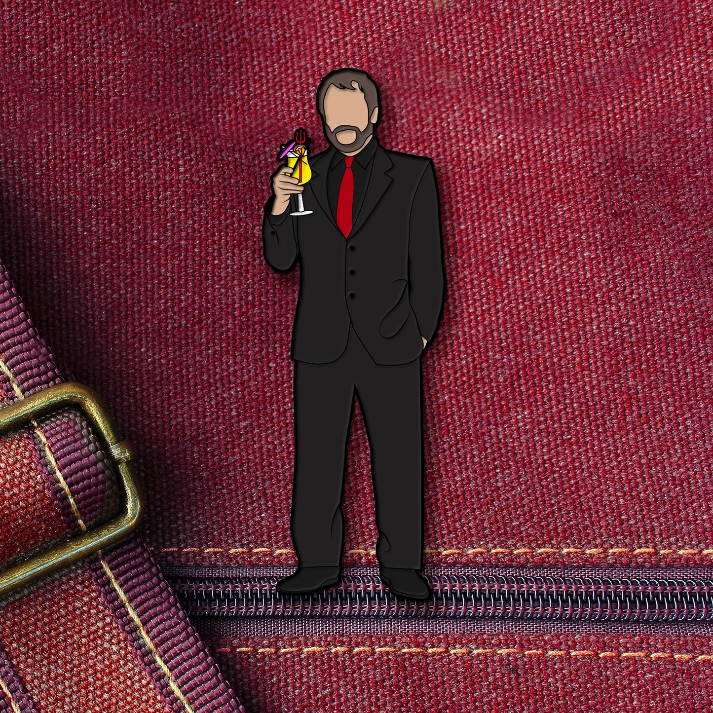 An enamel pin depicting Crowley holding a drink, attached to a maroon canvas bag.